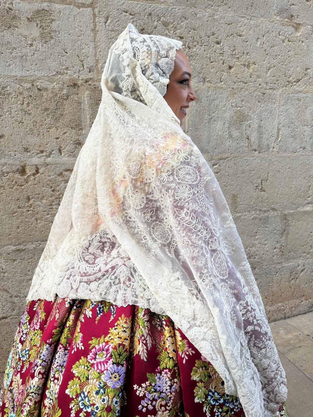 Mujer con indumentaria valenciana y mantilla clara junto a un muro de piedra